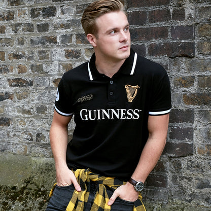 A man wearing a Guinness Black 59 White Stripe Polo Shirt from Guinness UK leans against a brick wall.