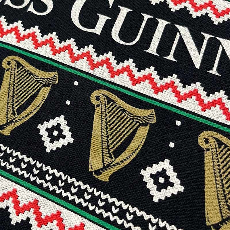 A close-up of the Guinness Festive Pints Polo Shirt showcases a stylish fabric featuring a red, white, and black geometric pattern with gold harps and partial "GUINN" text on a black background—ideal for the holiday fashion enthusiast seeking something unique from the Guinness brand.