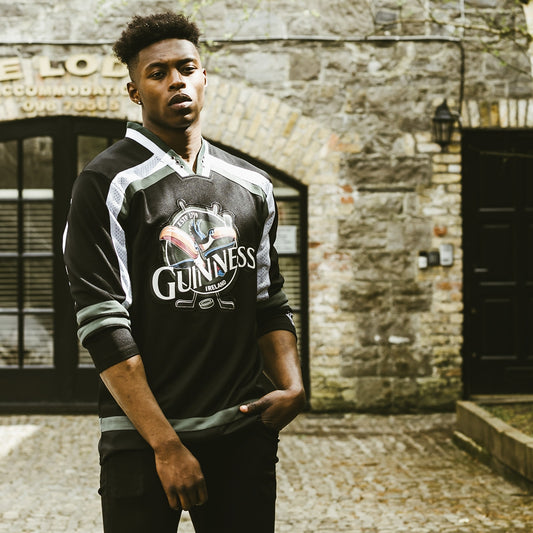 A person stands outdoors by a stone building, wearing a black and green Guinness Toucan Hockey Jersey from Guinness UK with an embroidered Guinness logo, looking at the camera.