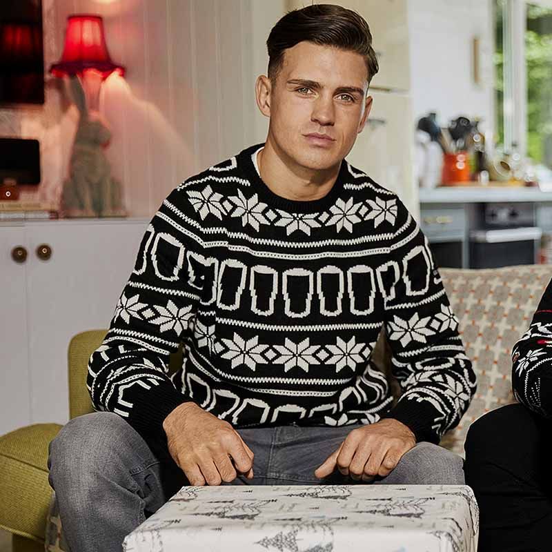 A man sits indoors wearing a cozy sweater radiating holiday spirit, dressed in the Guinness UK Festive Pints Christmas Jumper, adorned with a black and white pattern.