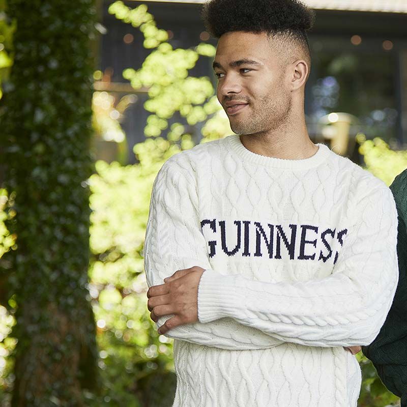 A person in a Guinness Cream Cable Knit Jumper stands outdoors amid lush greenery, highlighting the classic Aran style that makes it a true winter essential.