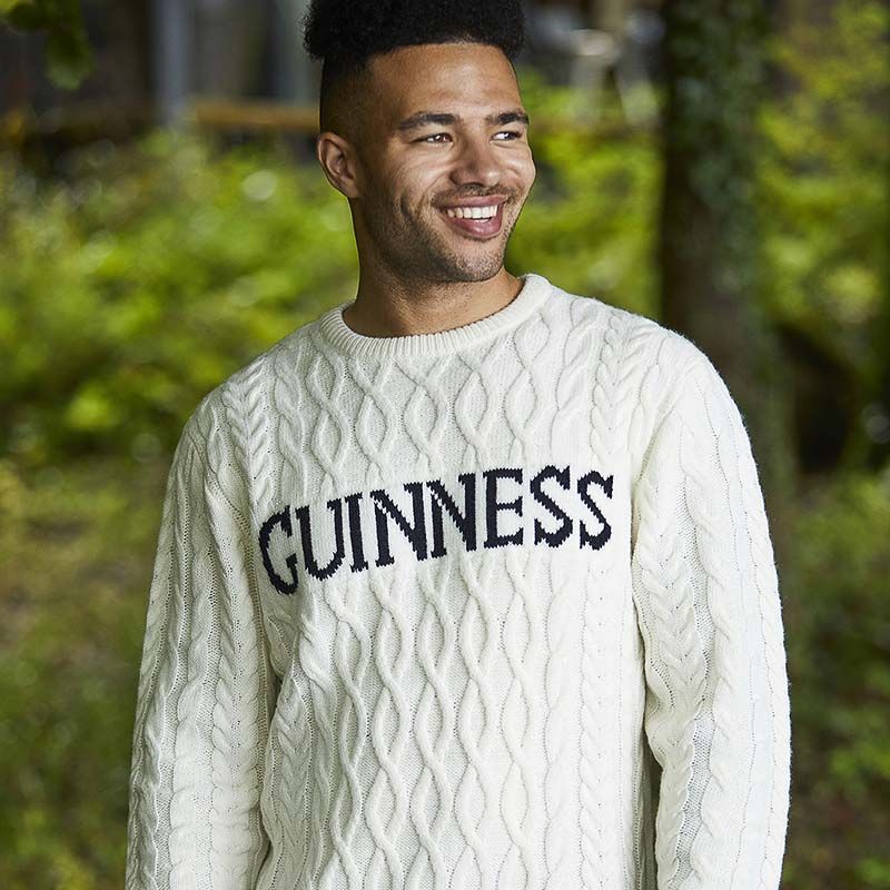 A man stands amidst lush greenery, wearing the Guinness Cream Cable Knit Jumper, complete with "GUINNESS" boldly displayed on the front—a true winter essential.