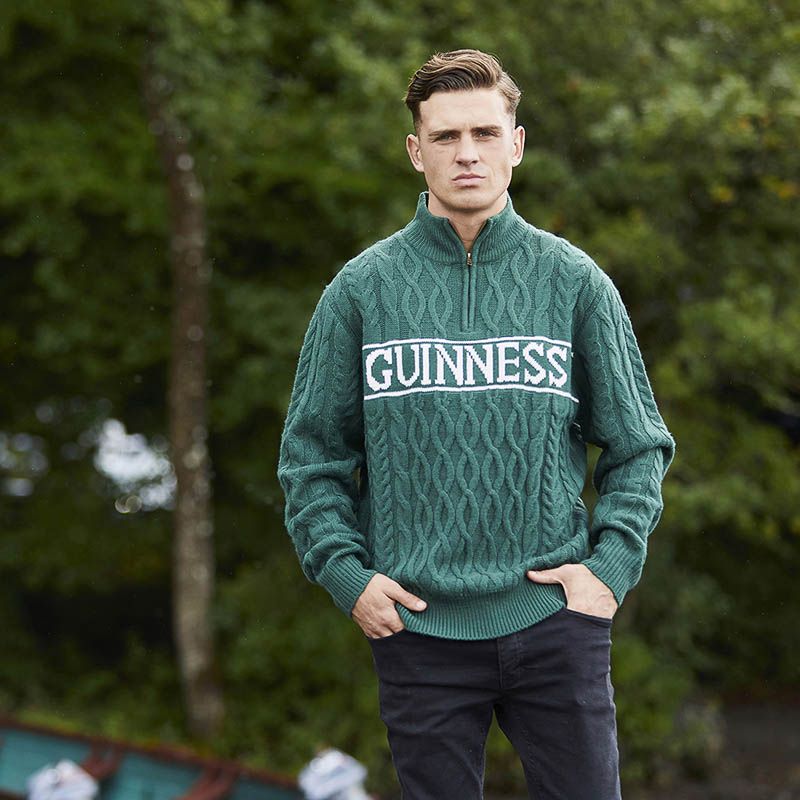 A person stands outdoors wearing the Guinness Green Cable Knit Quarter Zip Jumper with "Guinness" emblazoned on it. Trees are visible in the background.