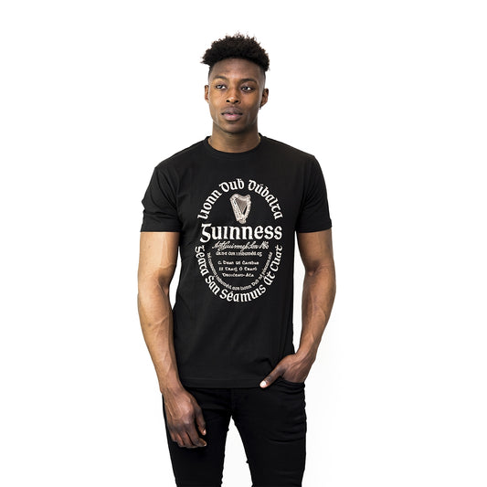Man stands against a white background wearing a Guinness UK Distressed Gaelic Label T-Shirt, an official black merchandise featuring white decorative text and graphics on the front.