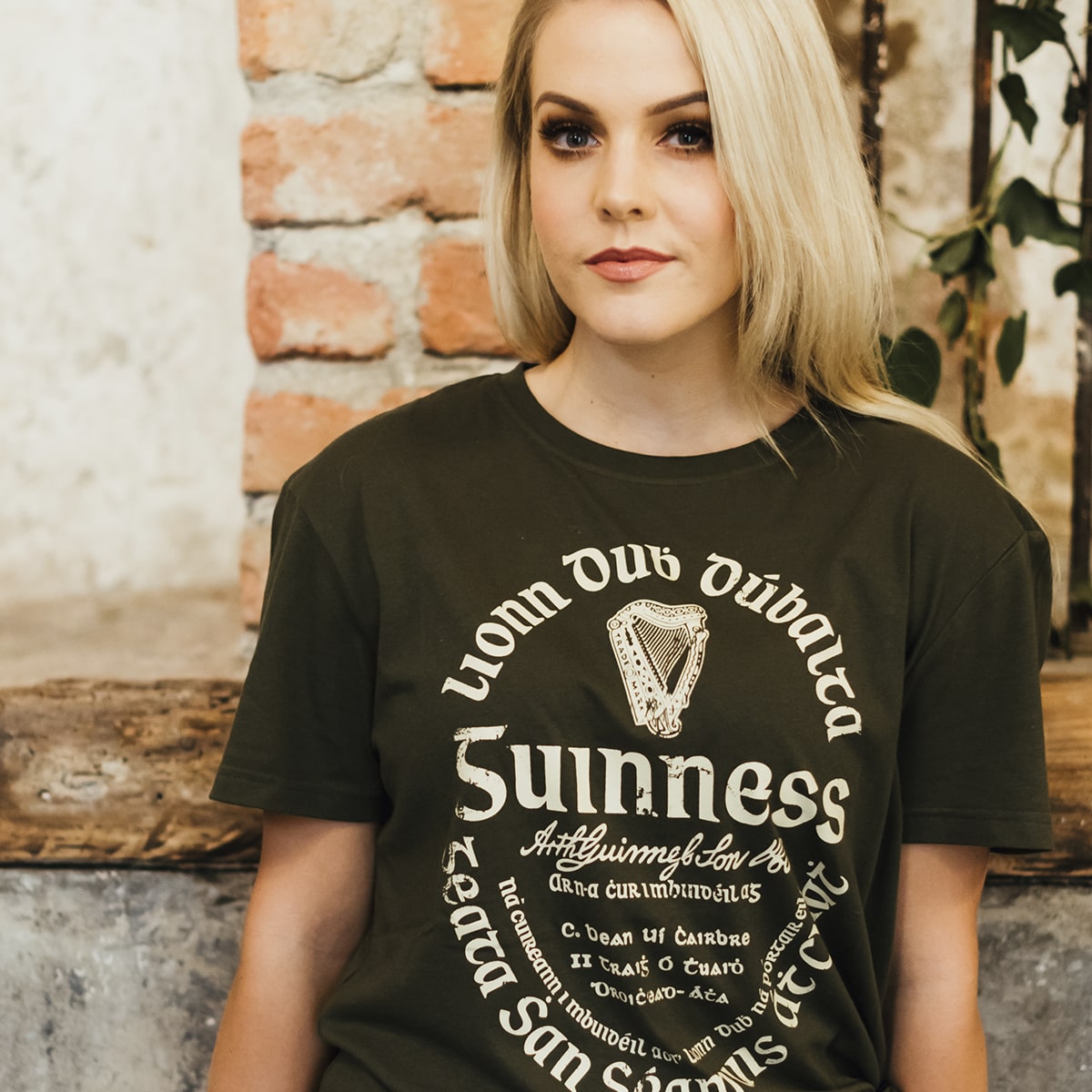 A woman with long blonde hair sits indoors by a brick wall, wearing the Guinness UK Khaki Green Gaelic Label Tee with cream lettering and the signature Gaelic label design.