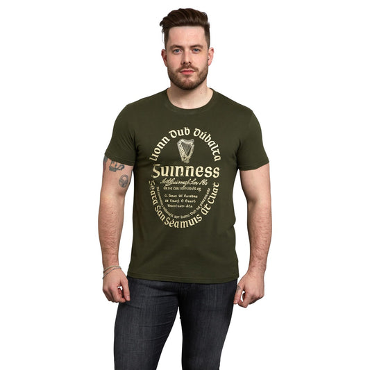 A man with short hair and a beard wears the Guinness UK Khaki Green Gaelic Label Tee, a unisex cotton t-shirt featuring the iconic white Guinness label, standing against a plain white background.