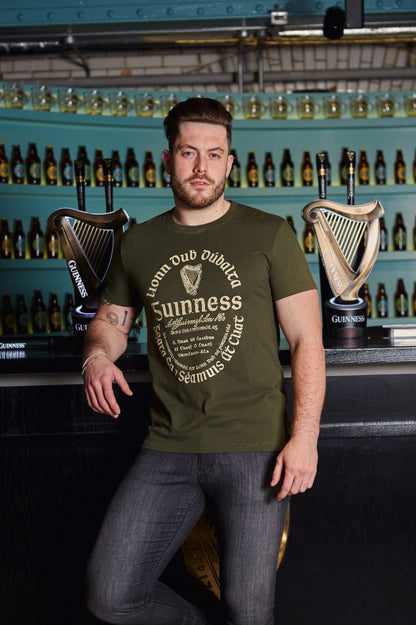 A man with short hair and a beard stands at a bar wearing a Guinness UK Khaki Green Gaelic Label Tee and jeans. Behind him, shelves of beer bottles and Guinness Trademark Label taps are visible.