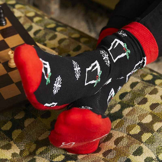 A person is sitting by a chessboard on a patterned cushion, sporting the festive Guinness Christmas Pint Socks Red and Black from the Guinness Webstore UK. These unique stocking fillers, with their black base adorned with white snowflakes and vibrant red heels, add a playful touch to the scene, perfect for any holiday gathering.