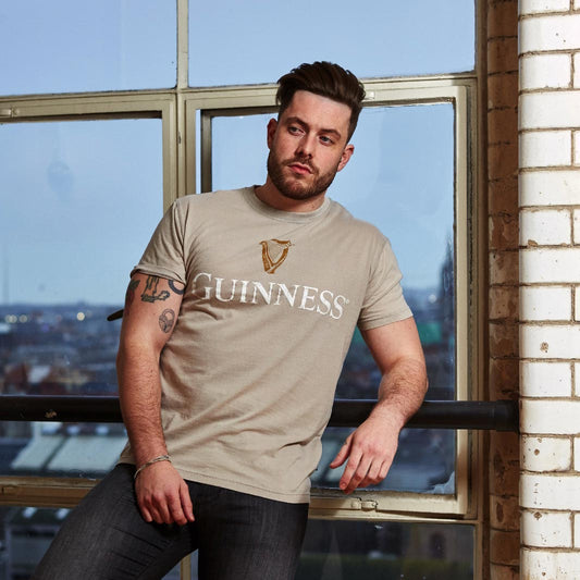 A man with short hair and a beard leans against a window ledge, wearing a beige Guinness Trademark Label T-Shirt from Guinness UK paired with dark jeans. The background shows an urban cityscape through large windows.