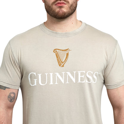 A person wearing a beige Guinness Trademark Label T-Shirt from Guinness UK, featuring the word "GUINNESS" and a harp logo on the front.