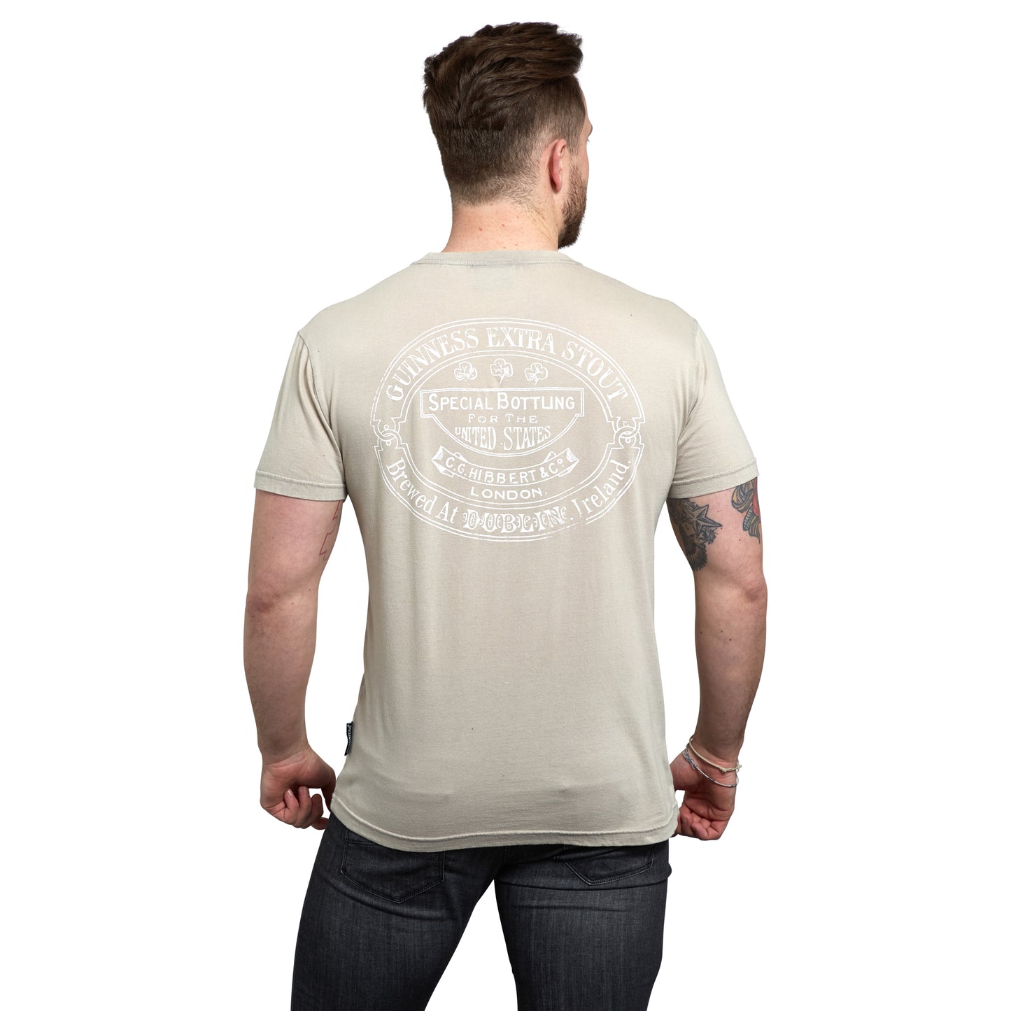 A man with a short haircut is wearing a GUINNESS TRADEMARK LABEL T-SHIRT BEIGE by Guinness UK, featuring a large vintage Guinness label and text on the back, paired with dark jeans. He is facing away from the camera.