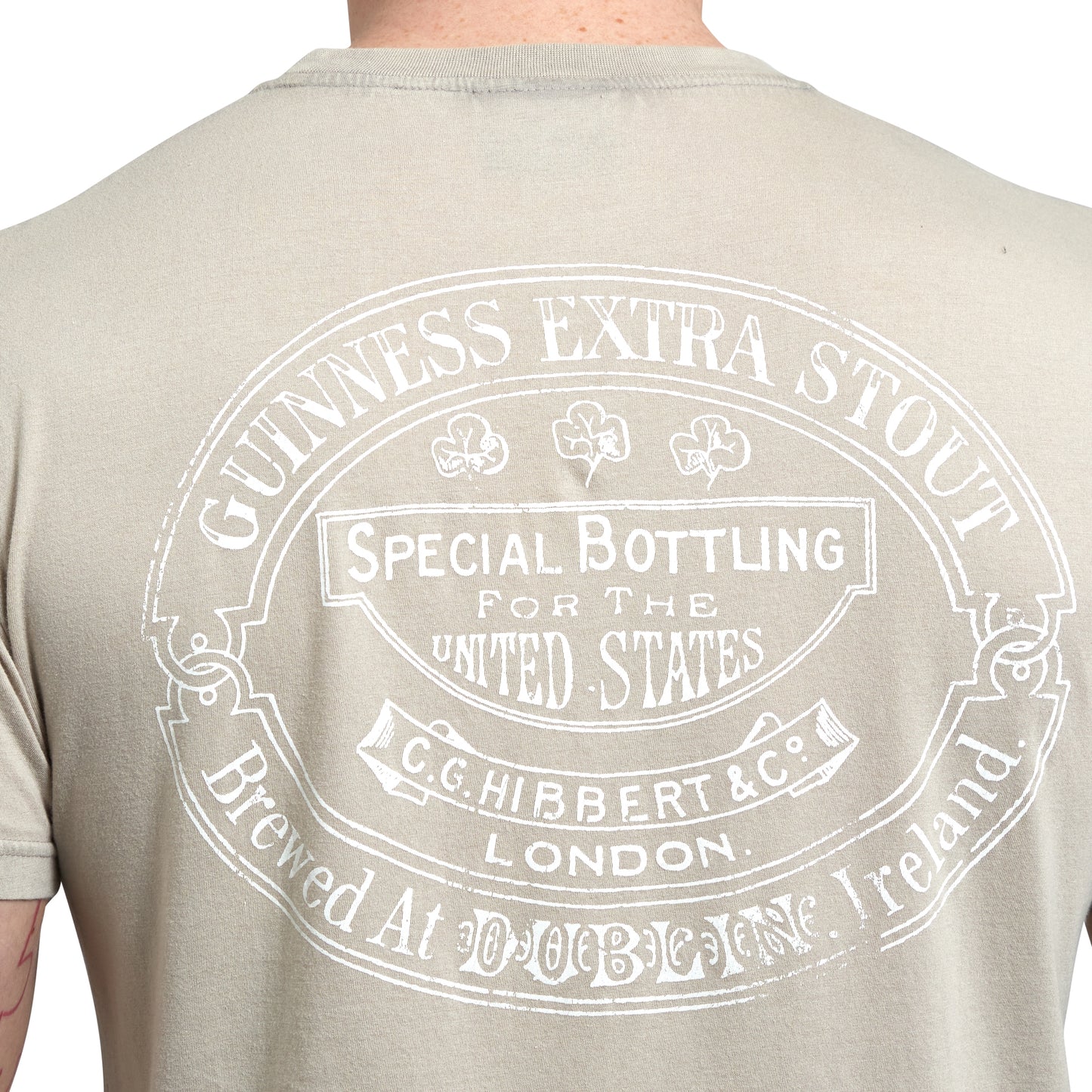 A person wearing a beige Guinness Trademark Label T-Shirt from Guinness UK, showcasing a white vintage-style design featuring text related to Guinness Extra Stout, including "Special Bottling for the United States" and "Brewed at Dublin Brewery, Ireland." The shirt proudly displays an iconic vintage Guinness label.