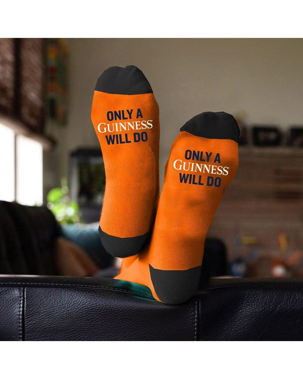 Feet adorned with Guinness Black & White Pint Socks, showcasing the iconic text "ONLY A GUINNESS WILL DO," rested comfortably on a black couch, bringing Irish charm to a cozy living room setting.