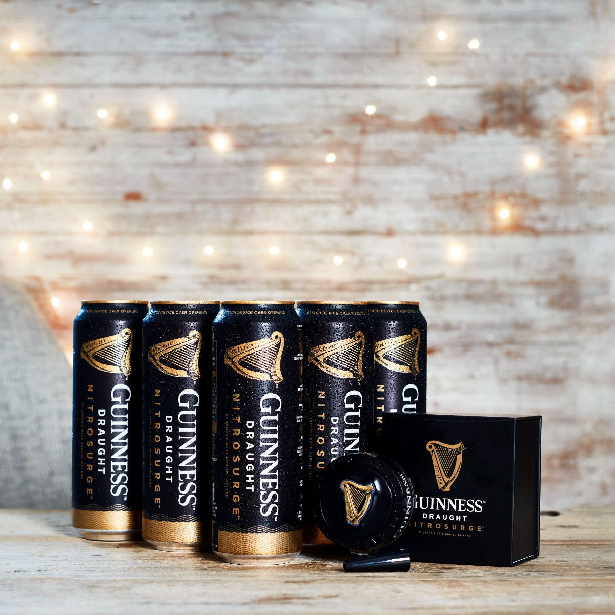 The Guinness Nitrosurge Gift Bundle, by Guinness UK, featuring cans of Guinness Draught and the innovative GUINNESS NITROSURGE device that creates nitrogen bubbles for the perfect pour, is showcased against a wooden background illuminated with string lights.