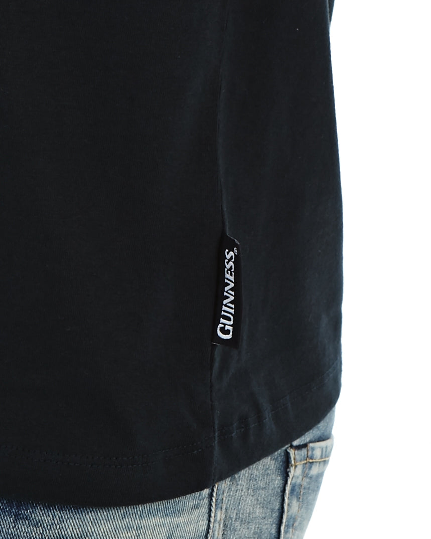 Close-up of the bottom edge of a black 100% cotton Guinness Gaelic Label Black Premium T-Shirt by Guinness UK, showing the tag on the side, paired with blue denim jeans.