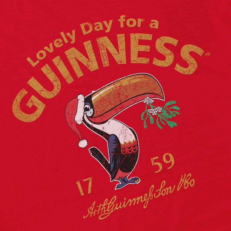 The red background of the Guinness Christmas Toucan T-Shirt Red displays a cartoon toucan wearing a Santa hat and holding mistletoe. The phrase "Lovely Day For A Guinness" is cheerfully written nearby, together with "1759 Arthur Guinness Son & Co." This festive tee from the Guinness Webstore UK adds a charming holiday touch.