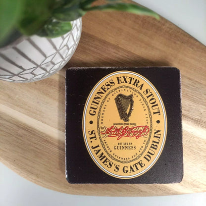 A Guinness Stone Effect Coaster - Extra Stout Label from the Guinness Webstore UK rests elegantly on a wooden surface, subtly shaded by a leafy plant in a stylish white pot.