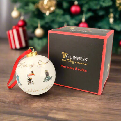 A festive Guinness Gilroy Ceramic Christmas Bauble with intricate designs is placed next to a black and red box from the Guinness brand's "Gilroy Collection," with the blurred image of a Christmas tree enhancing the seasonal charm in the background.