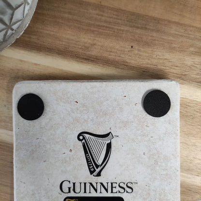 A close-up of the Guinness Stone Effect Coaster - Extra Stout Label from Guinness Webstore UK, featuring the iconic logo and two black circles on its surface. The coaster is placed on a wooden surface beside a patterned ceramic item.