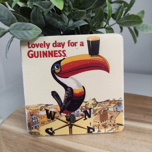 The Guinness Stone Effect Coaster - Gilroy Toucan from the Guinness Webstore UK features the classic design of a toucan balancing a pint on its beak next to a weather vane, accompanied by the phrase "Lovely day for a Guinness," making it an ideal gift for any fan of the iconic brew.