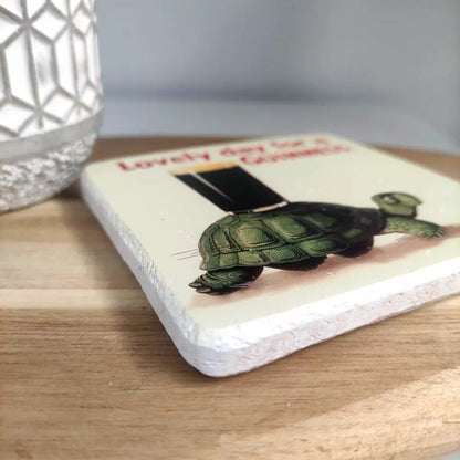 This delightful home accessory, the Guinness Stone Effect Coaster - Gilroy Tortoise from Guinness Webstore UK, showcases a ceramic coaster resting on a wooden base, adorned with an illustration of the Gilroy Tortoise carrying a stack of books. It’s perfect for adding character to any room and is as unique and charming as a collection of Guinness coasters.