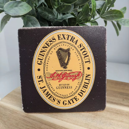 On a wooden surface beside a lush plant, the Guinness Stone Effect Coaster - Extra Stout Label from Guinness Webstore UK rests elegantly, showcasing its logo with a harp emblem.