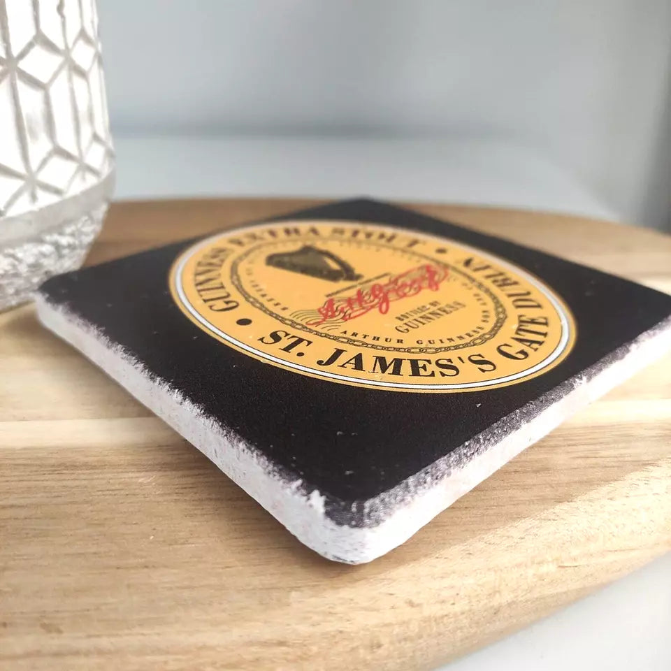 A Guinness Stone Effect Coaster, featuring the Extra Stout Label, is placed on a wooden surface next to a textured glass.