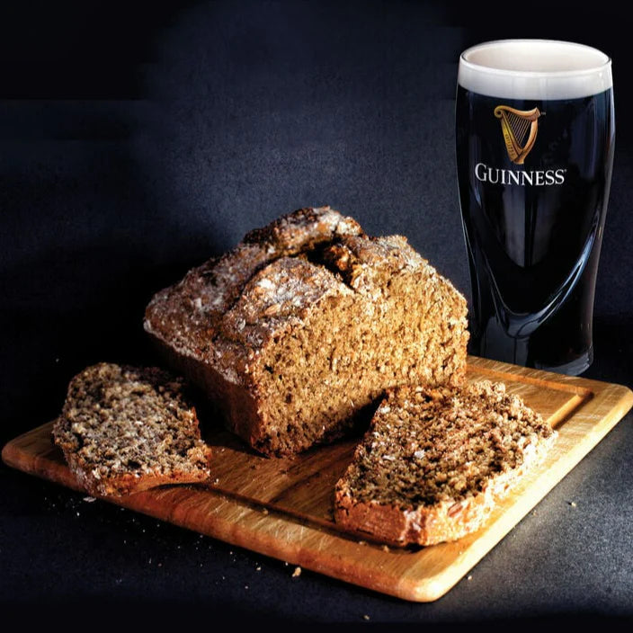 A loaf made with the Guinness Bread Kit from Guinness Webstore UK, featuring three slices on a wooden board, next to a pint glass of Guinness.