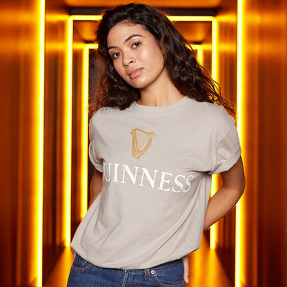 A woman with curly hair, wearing a beige GUINNESS TRADEMARK LABEL T-SHIRT from Guinness UK, stands in a brightly lit hallway with golden light panels.