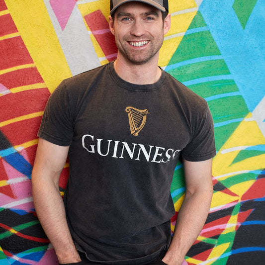 A man wearing a Guinness Distressed Trademark Label T-Shirt leaning against a colorful wall.