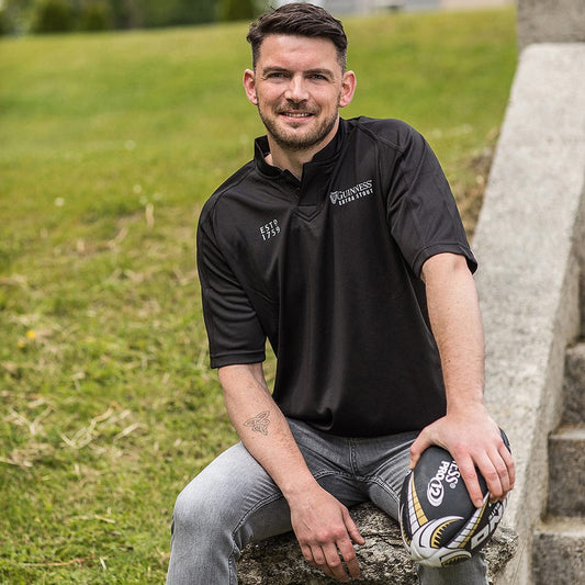 A man sitting on some steps with a rugby ball, wearing a Guinness UK All Black Rugby Jersey.